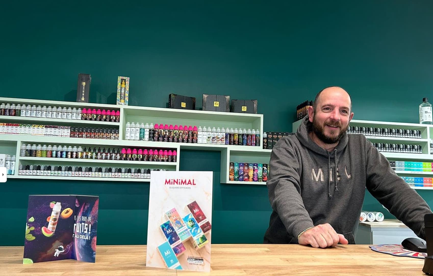 Fred, souriant derrière le comptoir de la boutique, devant le rayon d’e-liquides.