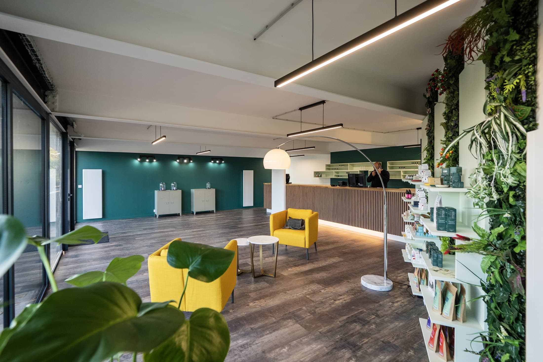 La salle de la boutique vue depuis l’entrée : fauteuils jaunes, guéridons, mur végétal et comptoir en bois.